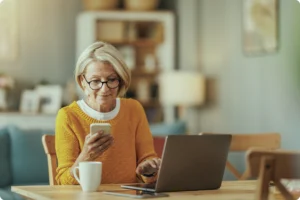 Older woman using a smartphone and laptop at a table while accessing a digital service.
